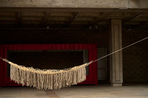 Une œuvre d'art suspendue ressemblant à un tapis volant, faite de pailles de roseaux et de fil de lin brut, exposée dans un espace intérieur avec un fond rouge.
